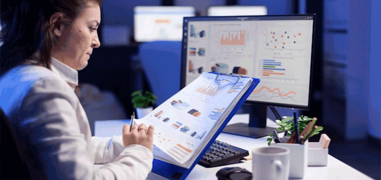 Mulher sentada numa mesa de escritório enquanto analisa papel com vários gráficos, na mesa encontra-se um notebook com a tela também mostrando vários gráficos