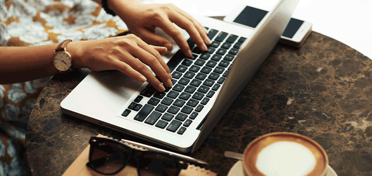 Uma mulher digitando em um laptop ao lado de uma xícara de café