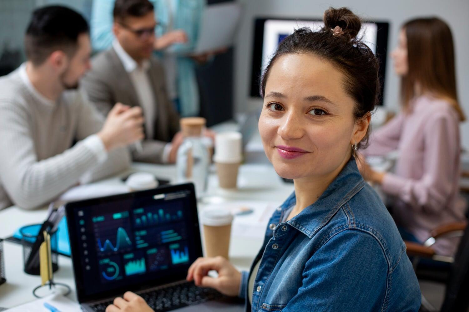 Profissional sorridente com laptop exibindo dashboards em ambiente colaborativo de equipe