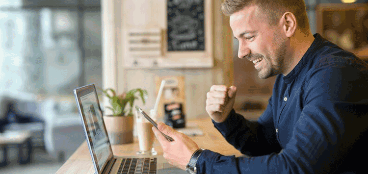 Homem sentado em uma mesa de frente para um notebook, enquanto segura um celular e faz gestão de comemoração