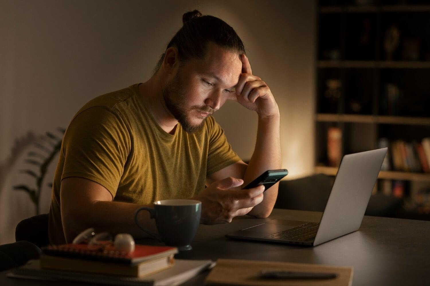 Homem pensativo à noite olhando o celular em frente ao notebook